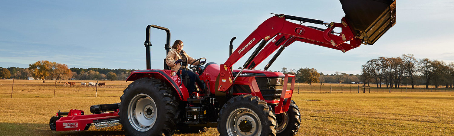 A woman on a ranch, driving a red Mahindra® tractor with the bucket up.